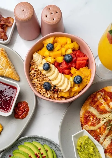 Brunch table with a fruit and granola bowl topped with banana and blueberries, pastries, jam and a pizza